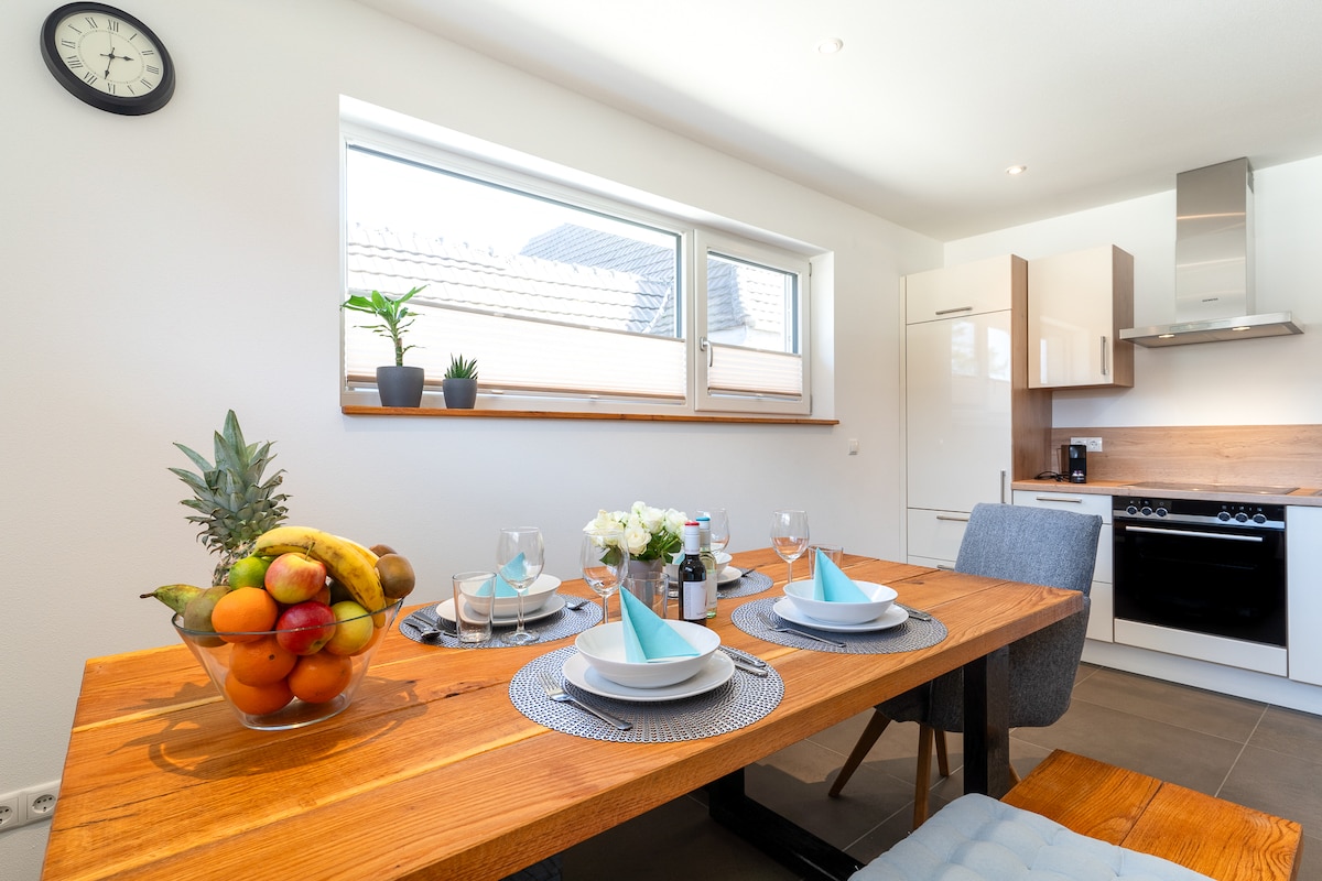 A sunlit dining area features a wooden table set for a meal. A bowl of fresh fruit is displayed at the center, accompanied by elegant place settings. Large windows provide natural light, while the modern kitchen is partially visible in the background.