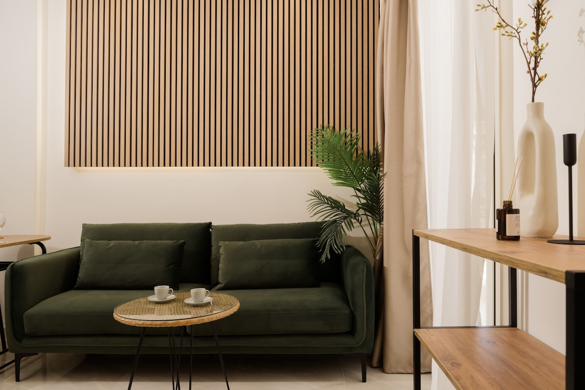 A cozy living area features a green sofa positioned against a backdrop of wooden slats. Two small cups rest on a round rattan coffee table. A potted plant adds a touch of greenery, while soft curtains frame the window, enhancing the inviting space.