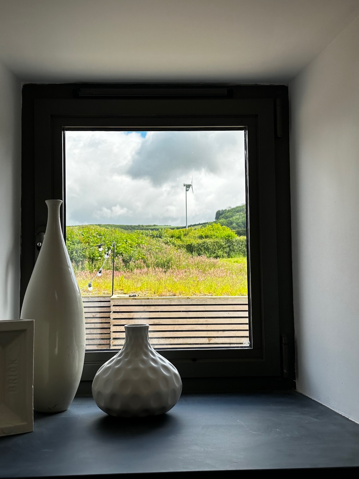 A window frame is visible, offering a view of lush green fields under a partly cloudy sky. Two decorative vases, one tall and white and the other round and textured, are placed on a dark surface in front of the window.