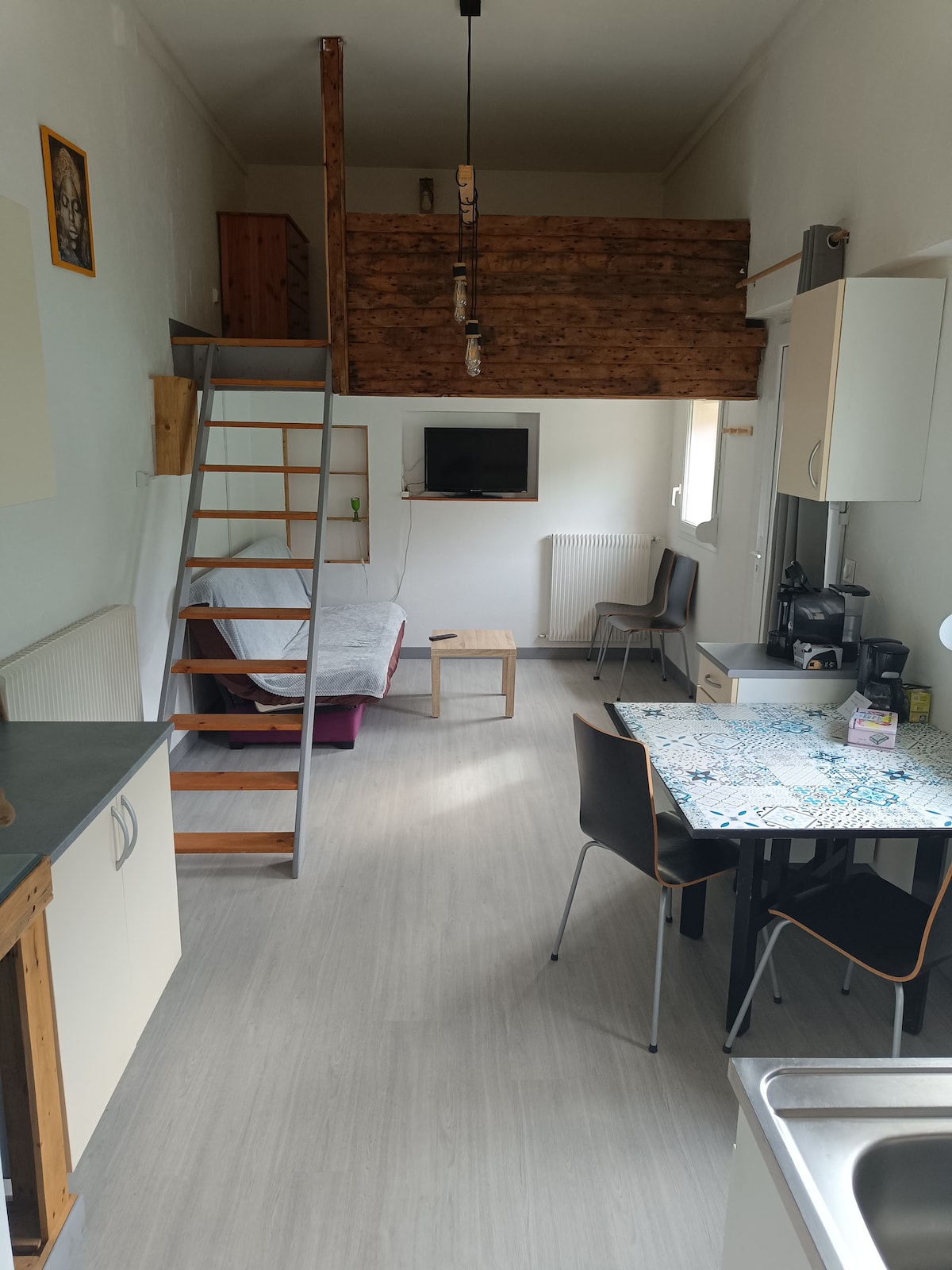 The studio features a compact layout with a wooden staircase leading to the mezzanine sleeping area. A television is mounted on the wall, while a small coffee table and couch are positioned in the living space. The dining table is set near the kitchen area, offering a functional setup.