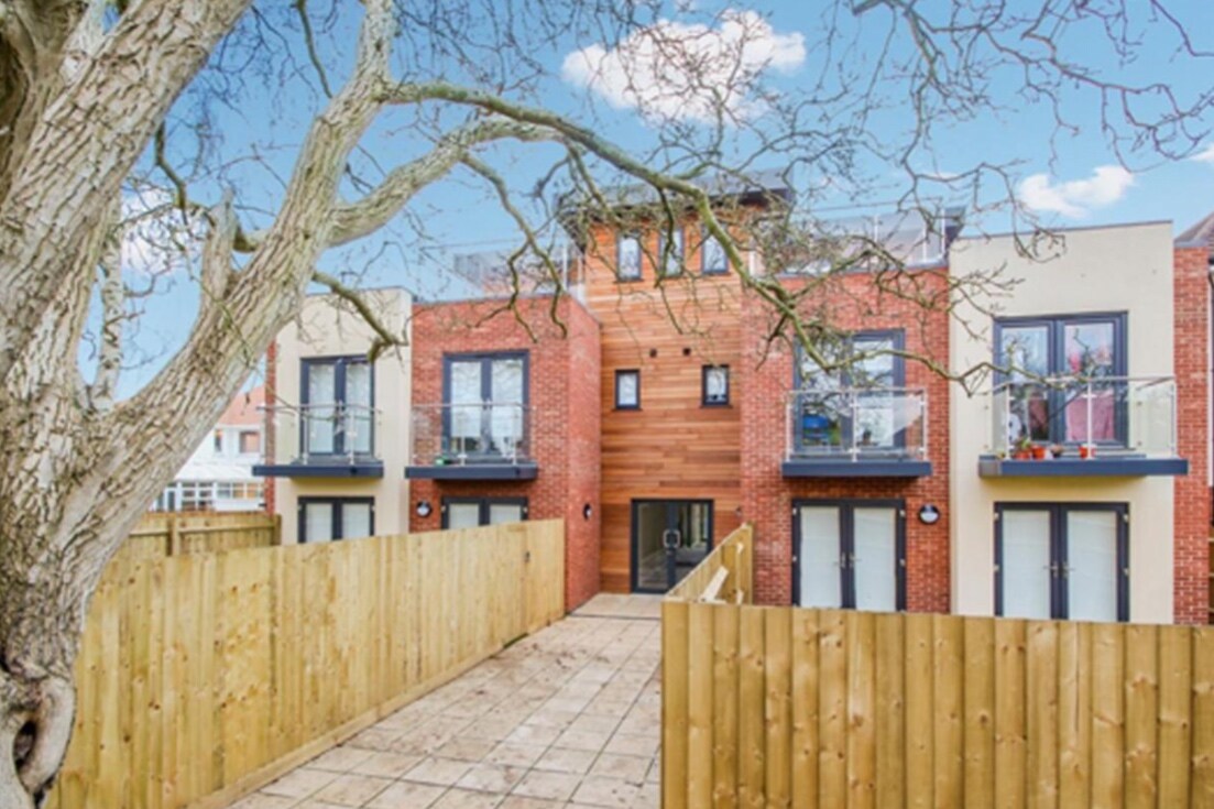 A modern building with a mix of brick and wood accents is framed by a leafless tree. The entrance is accessed via a paved pathway, flanked by a wooden fence. Three balconies extend from the upper level, adding charm to the contemporary design.