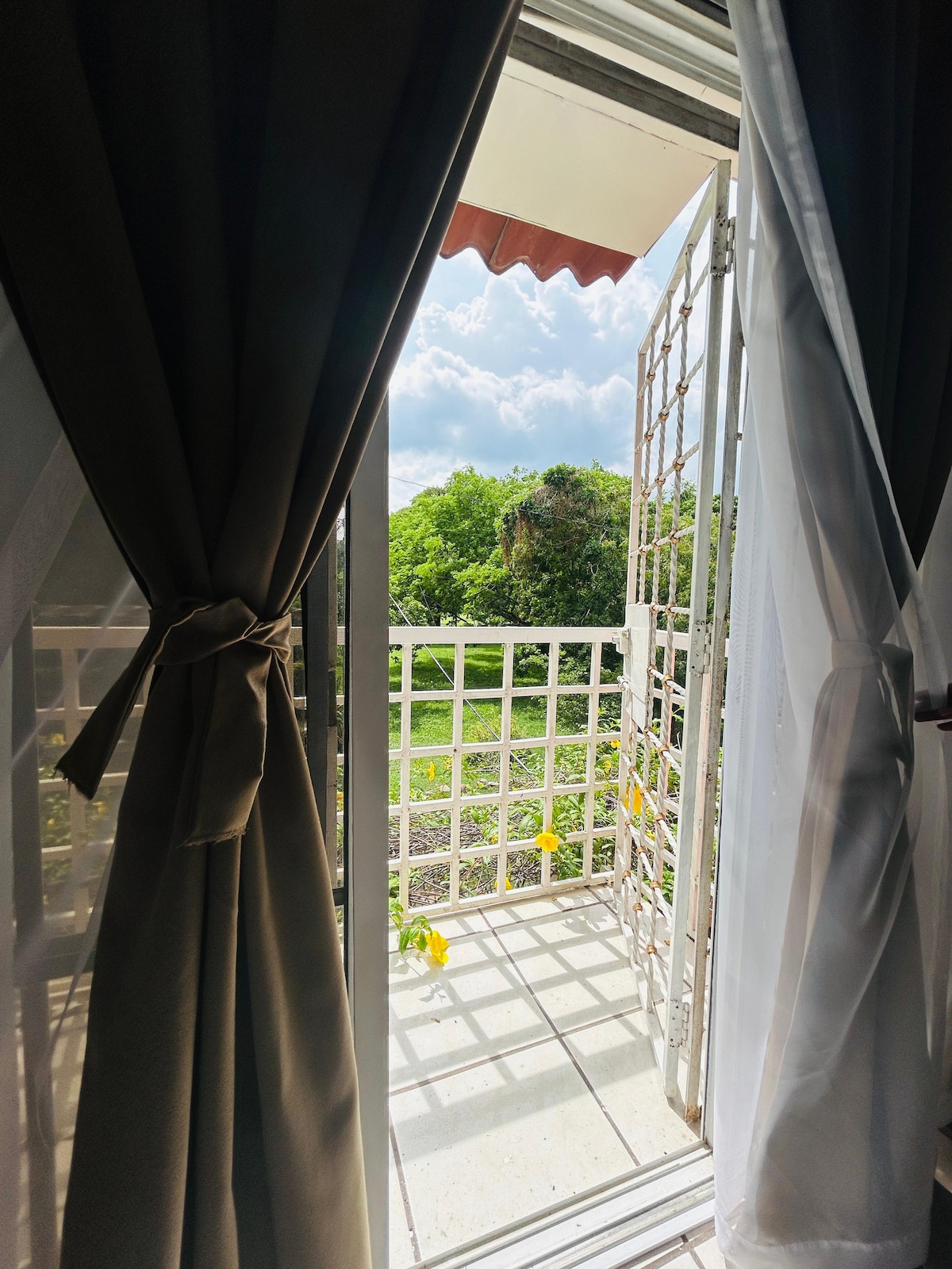 A balcony door is partially open, revealing a green outdoor space bathed in natural light. The view includes lush trees and a well-maintained garden area. Sheer white and solid dark curtains frame the scene, enhancing the tranquil ambiance of the setting.