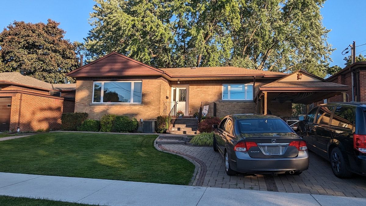 A detached bungalow is presented with a brick exterior and a front walkway leading to the main entrance. Lush green lawns frame the property, complemented by mature trees. A driveway is visible, accommodating multiple vehicles, further indicating accessibility and space.