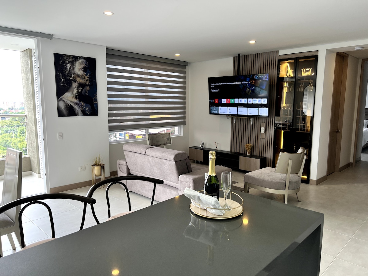 An open living area features modern furnishings, including a gray sofa and stylish chairs. A large television is mounted on the wall, adjacent to floor-to-ceiling windows that allow natural light to fill the space. An elegant dining bar is visible in the foreground.