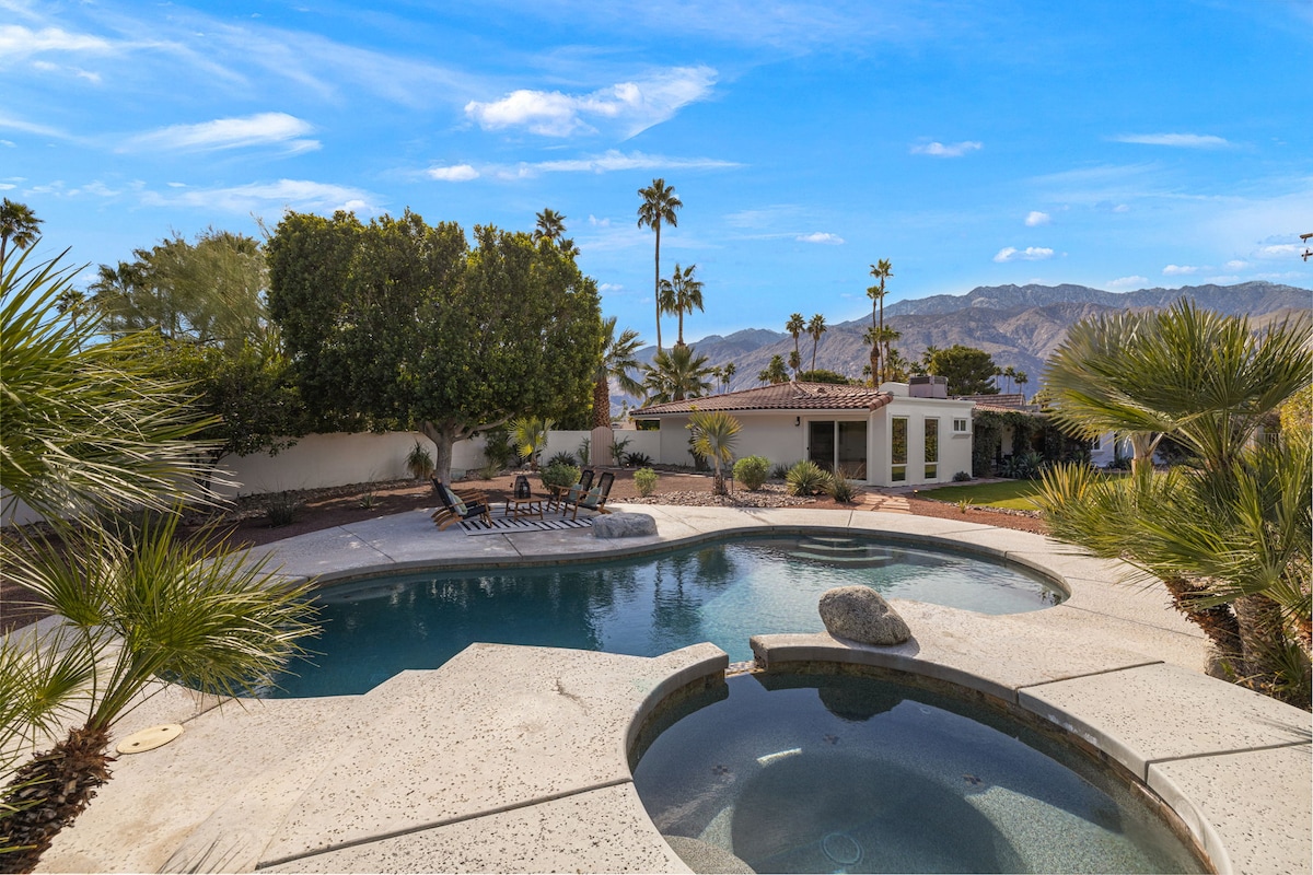 An inviting backyard showcases a sparkling pool and a hot tub, surrounded by desert landscaping. Lush palms and vibrant greenery frame the area, while mountain views stand in the background under a clear blue sky. Comfortable lounge chairs are positioned by the pool.