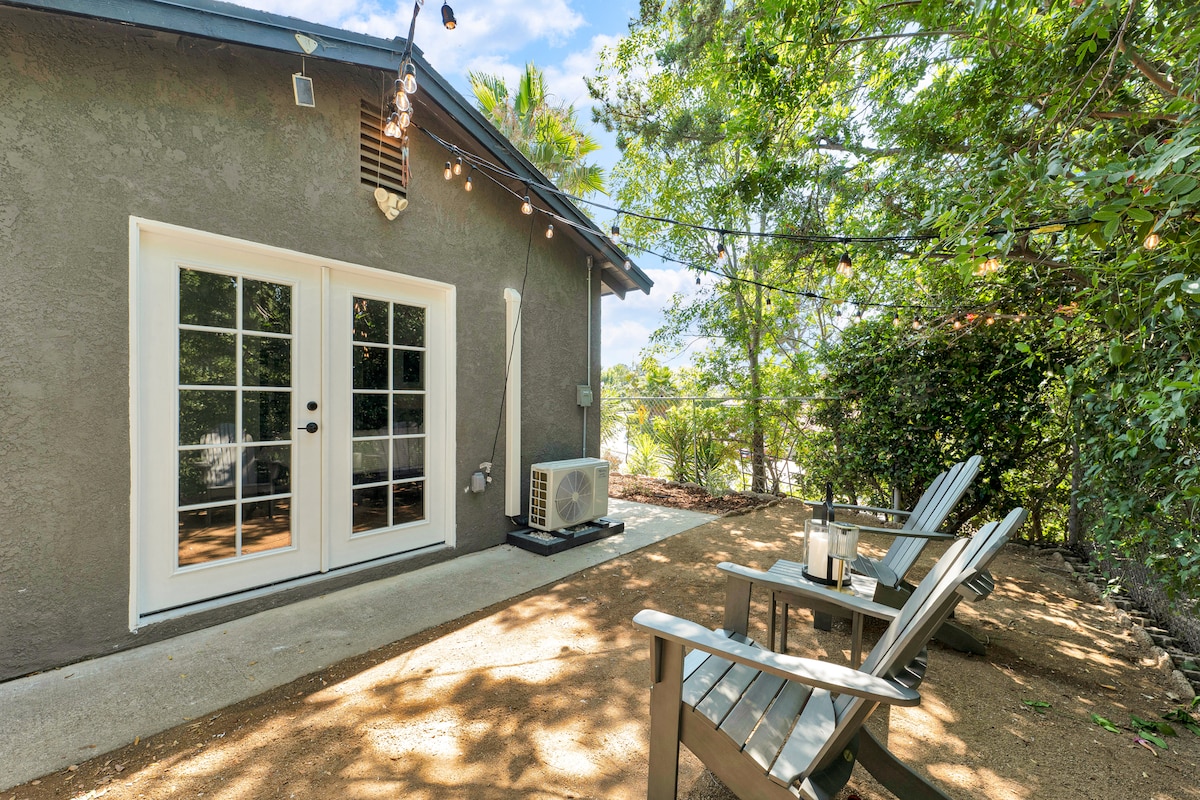 A private outdoor area features two wooden chairs positioned on a sandy surface, surrounded by lush greenery. A set of decorative string lights hangs above, enhancing the inviting space, while double glass doors lead into the guest house, providing access to the interior.