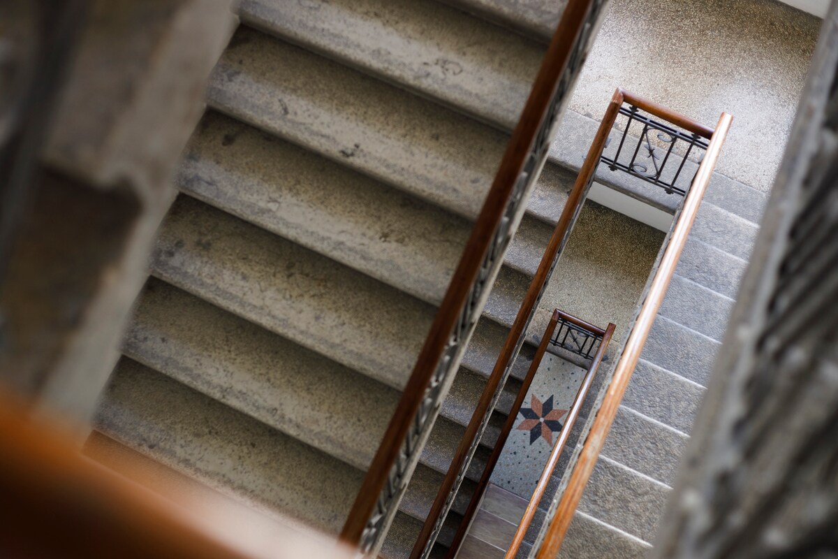 A view of a staircase featuring stone steps and wooden railings. Decorative elements are visible on the landing, including a patterned tile designed with star motifs. Natural light illuminates the staircase, creating a welcoming atmosphere in this historic building.