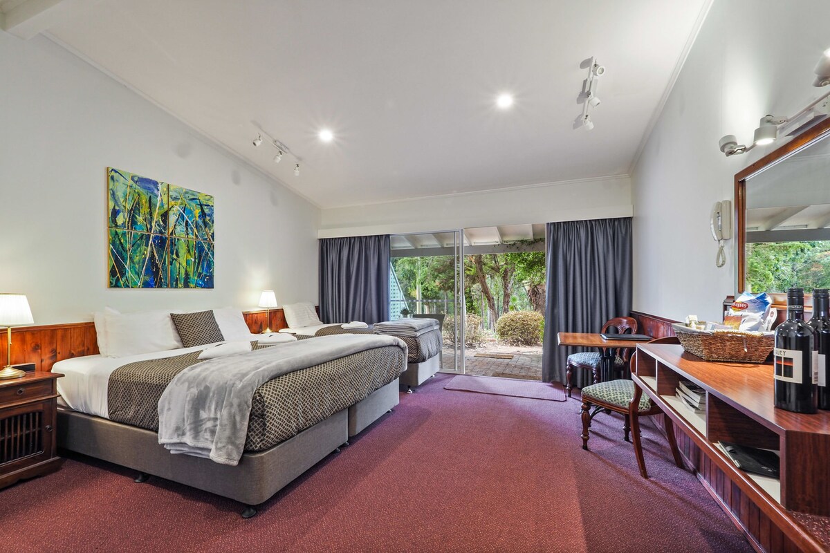 The studio suite features two spacious beds with neutral bedding, complemented by bedside lamps. A desk with a mirror and refreshments is positioned nearby. Large glass doors open to a terrace, offering views of the surrounding greenery. Natural light fills the room, enhancing the inviting atmosphere.