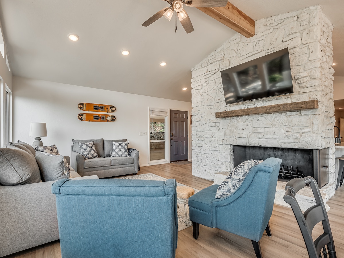 A cozy living area is highlighted by a stone fireplace and mounted television. Two blue armchairs and a gray sofa provide seating, while decorative pillows add comfort. Natural light fills the space through large windows, complemented by a ceiling fan above for airflow.