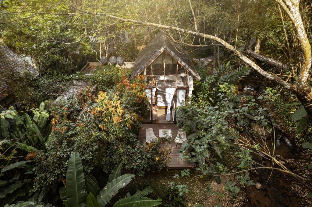 A charming villa is nestled among lush greenery, featuring an outdoor deck with seating. The roof is thatched, blending harmoniously with the surrounding natural landscape. Tall trees and vibrant foliage create a serene atmosphere, while a small stream can be seen nearby.