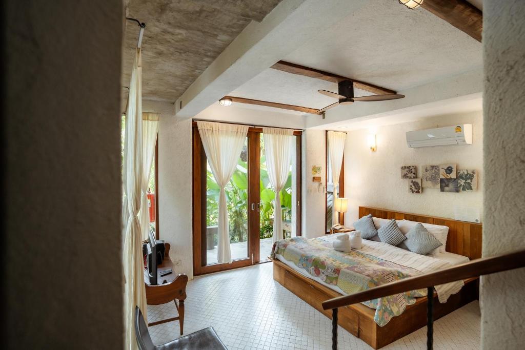 A bright and airy villa room features large glass doors that open to a private balcony, surrounded by lush greenery. A spacious bed with decorative pillows is set against a wooden bedframe. A small seating area is present, along with a ceiling fan and artwork on the walls.