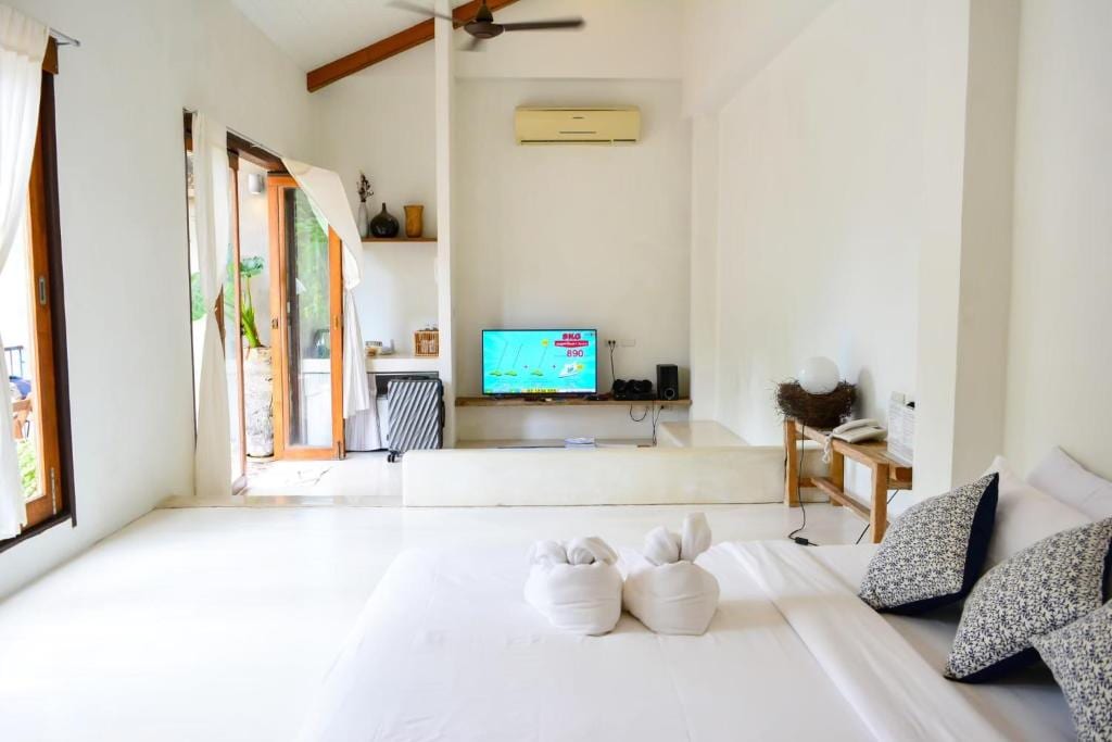 A bright and airy villa interior features a spacious bed adorned with decorative towels resembling swans. A flat-screen TV is positioned opposite the bed, alongside a minimalistic entertainment area. Large windows provide natural light and access to a private balcony, enhancing the open layout.
