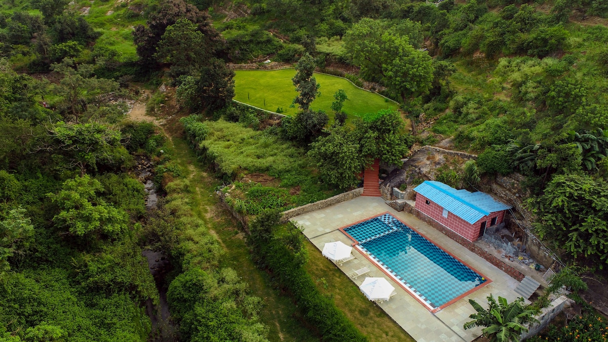 An aerial view showcases a serene landscape featuring a swimming pool bordered by a patio and three white lounge chairs. Lush greenery surrounds the area, complemented by a grassy field in the background and a modest building with a blue roof nearby.