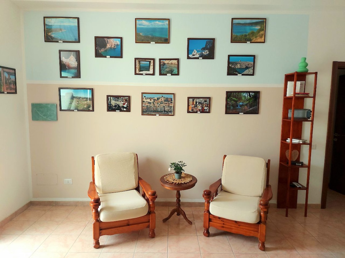 A welcoming sitting area features two wooden armchairs complemented by a small round table. The wall is adorned with a variety of framed photographs depicting scenic landscapes. A bookshelf stands nearby, showcasing decorative items and providing an inviting atmosphere for relaxation.