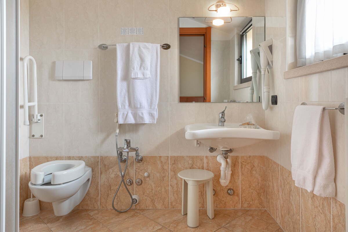 A well-appointed bathroom is shown, featuring a toilet, sink, and a shower. Handrails are installed for accessibility. Soft towels hang neatly on hooks, and a mirror is positioned above the sink, enhancing the space with natural light from a nearby window.