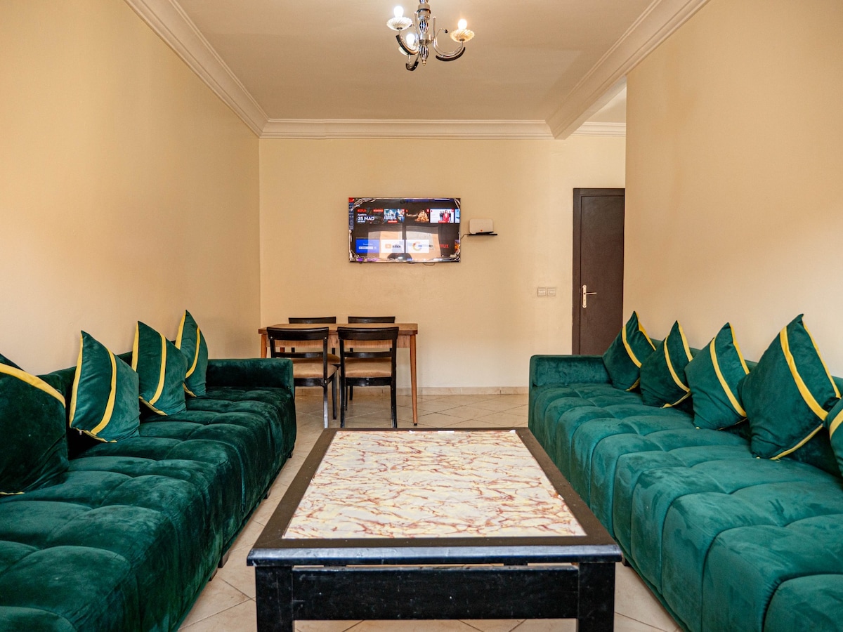 A spacious living room is presented, featuring two long green sofas adorned with yellow cushions. A coffee table with a patterned surface centers the space. A flat-screen TV is mounted on the wall, with a dining table and chairs positioned in the background.