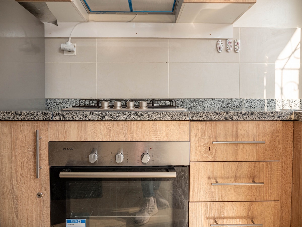 A modern kitchen area features light wood cabinetry complemented by a granite countertop. A stainless steel oven sits below a five-burner gas cooktop, offering ample space for meal preparation. Clean lines and a minimalist design contribute to the functional layout.