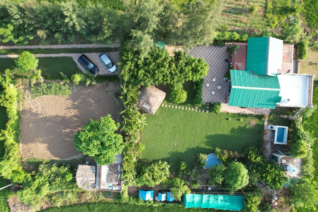 An aerial view captures a lush green landscape featuring a combination of cultivated land, trees, and a well-maintained garden. A thatched-roof structure is visible, along with two parked vehicles and a spacious area laid with paved stones, providing an inviting outdoor setting.