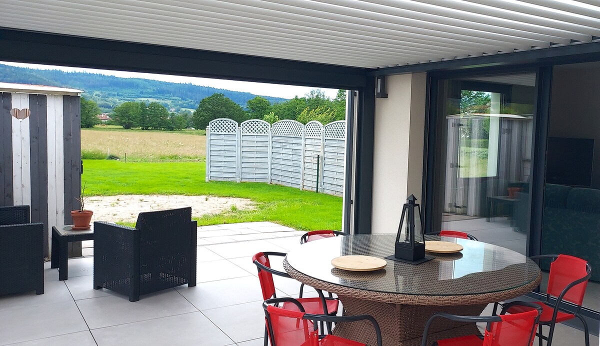 An outdoor terrace is furnished with a round dining table and red chairs, providing a space for meals. Nearby, a cozy seating area with dark wicker chairs complements the view of the green landscape and scenic backdrop.