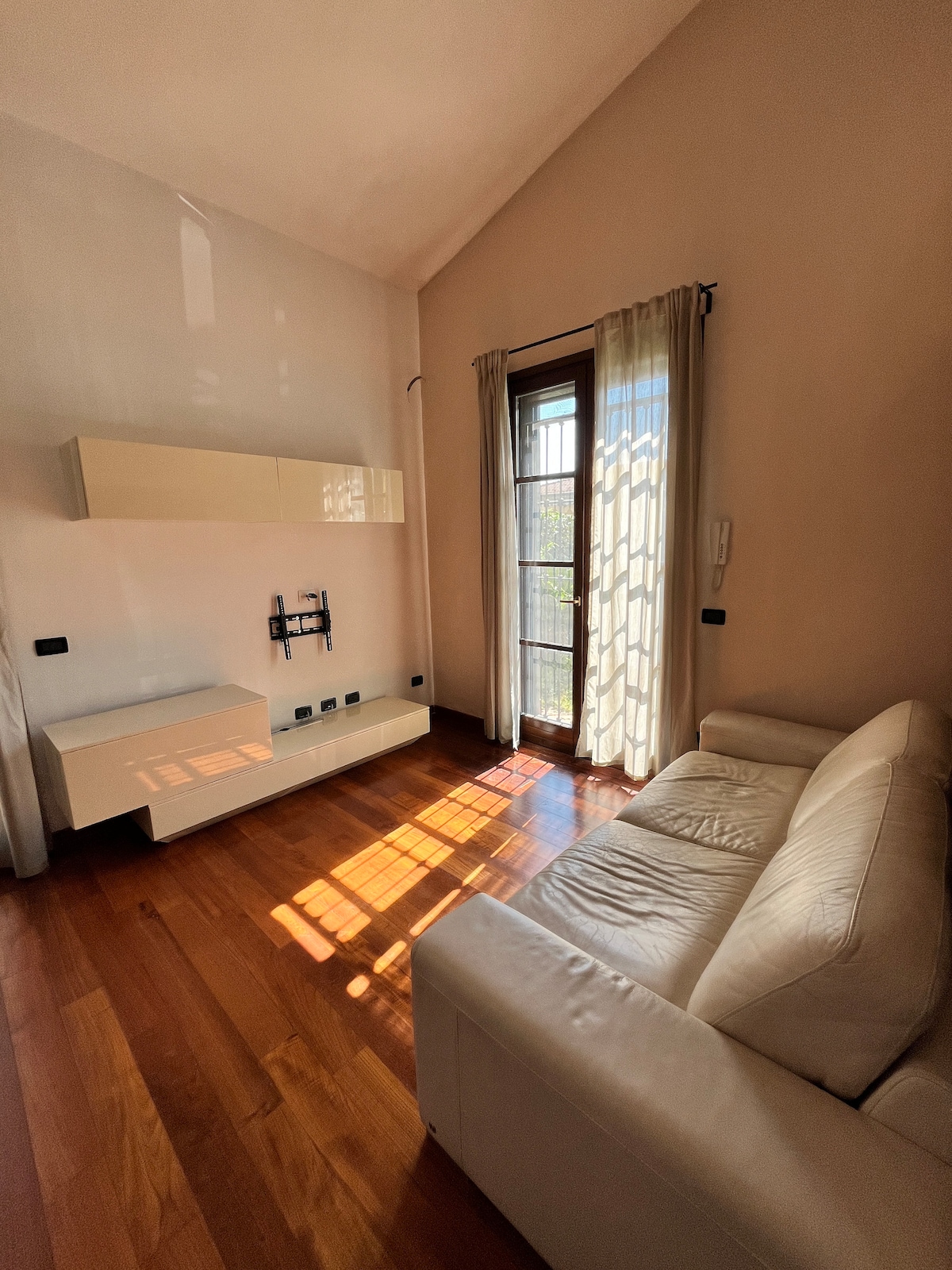 A living space is highlighted by a white sofa positioned on polished wooden flooring. Natural light filters through a set of curtains, casting patterns on the floor. A wall-mounted entertainment unit is present, along with a balcony door leading outside.