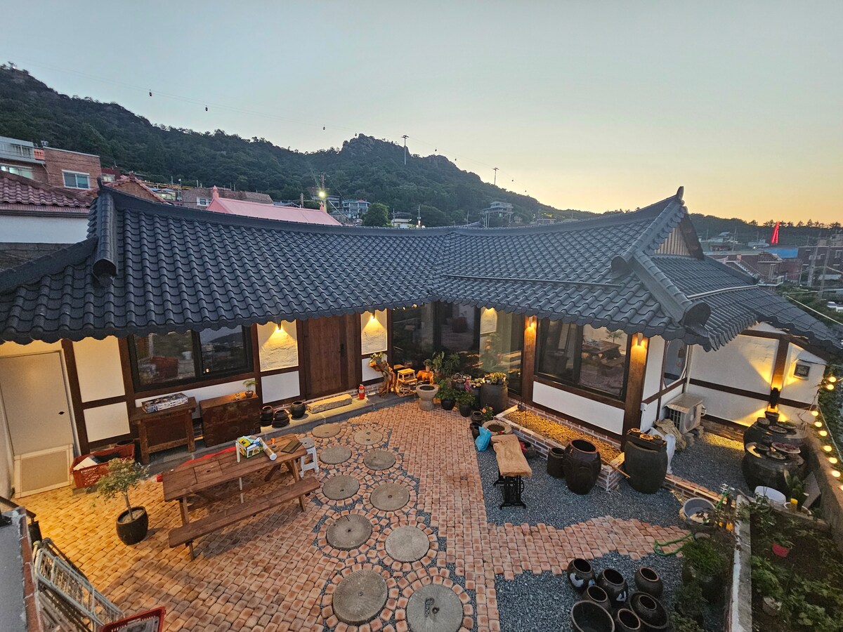 An outdoor area features a stone and brick pathway surrounded by various potted plants and traditional pots. A large wooden table provides ample seating, while the unique eaves of the structure are highlighted against the backdrop of a sunset sky.