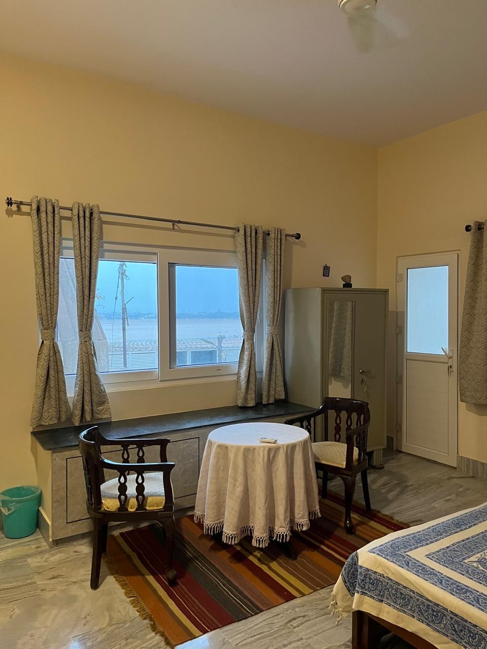 A well-lit room features two wooden chairs surrounding a round table with a lace tablecloth. A large window offers views of the river, while a wardrobe and a door are also visible. Soft linens adorn the spacious bed, and the floor is covered in a striped rug.