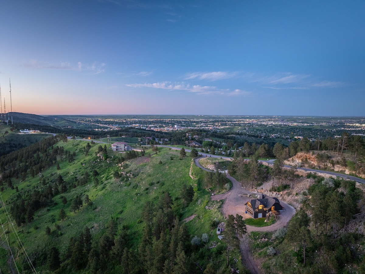Aerial view of a house situated on a hillside, showcasing surrounding lush greenery and rolling terrain. The expansive skyline of Rapid City is visible in the distance, bathed in soft evening light, while winding roads lead through the scenic landscape.
