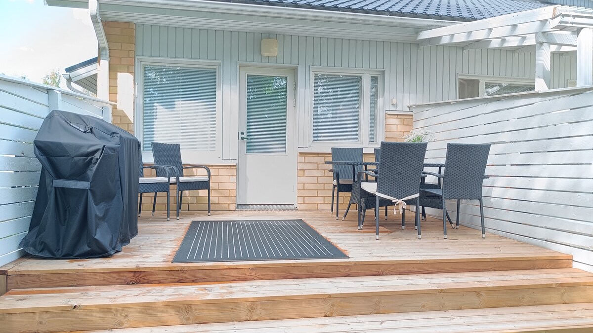 An outdoor terrace is featured, showcasing a wooden deck with a black gas grill and six black patio chairs surrounding a glass table. Natural light enhances the space, framed by white railing and a light-colored exterior.