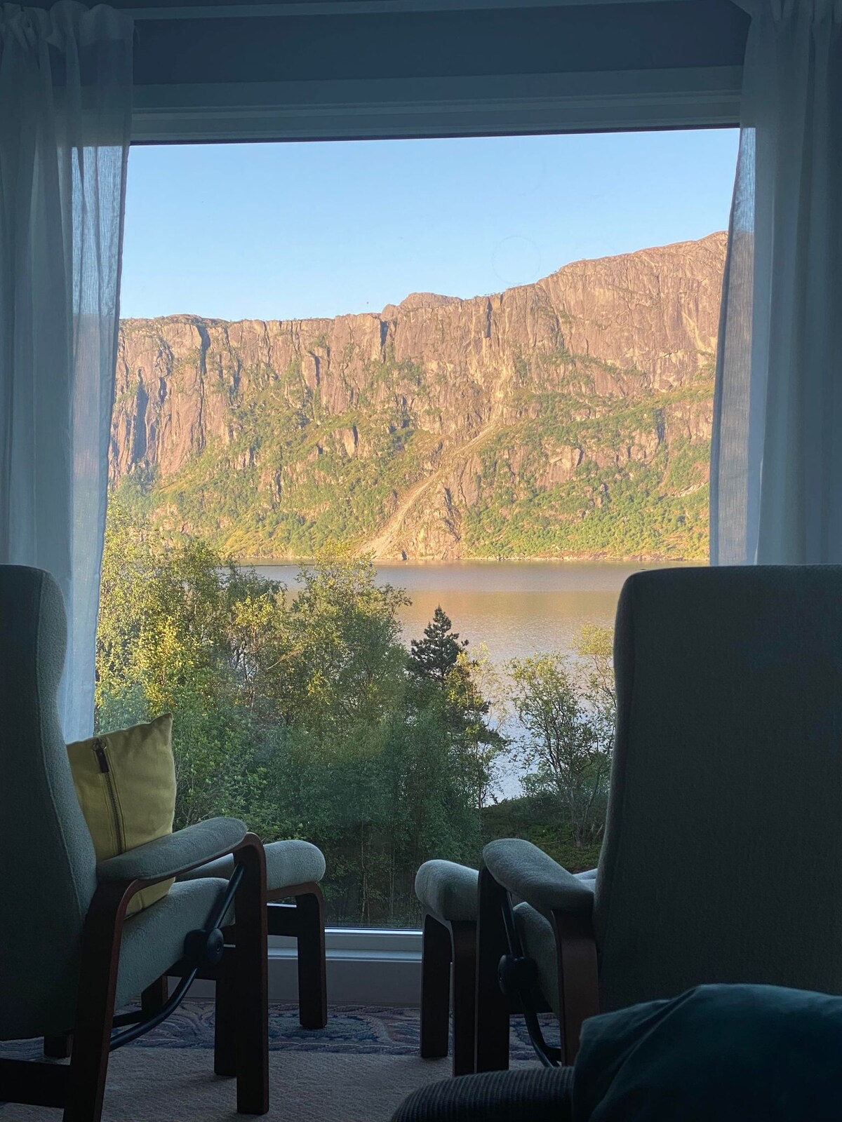 Two comfortable chairs are positioned by a large window, offering a view of a calm fjord framed by steep cliffs and lush greenery. Natural light filters through sheer curtains, highlighting the serene landscape outside.