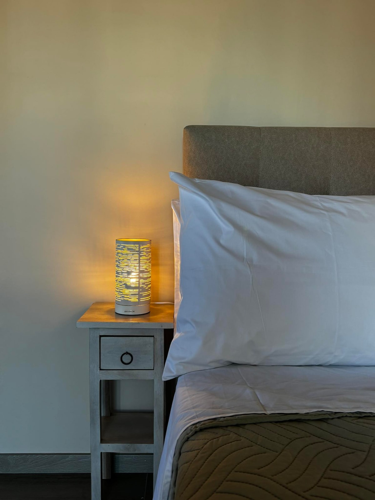 A spacious bed is positioned against a soft-colored wall, complemented by a bedside table. A lamp with a patterned design emits a warm light, enhancing the serene atmosphere of the sleeping area. Crisp white bedding and a neutral-toned quilt are visible.