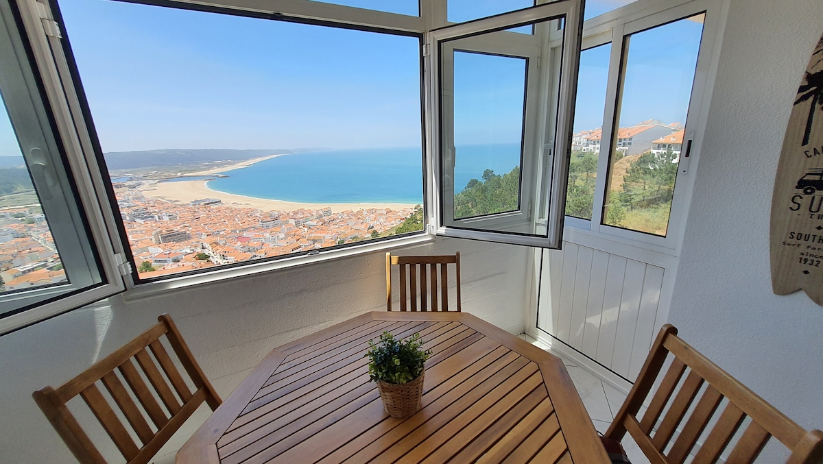 A bright dining area features a wooden table surrounded by four chairs, set against large windows offering expansive views of the Atlantic Ocean and Nazaré town. The ambiance is enhanced by natural light streaming through, creating a relaxed and inviting space.