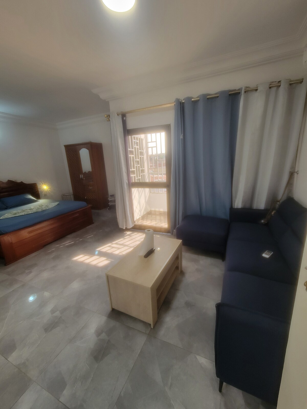 A spacious room features a wooden bed with light bedding. A dark blue sofa is arranged next to a small coffee table, while natural light filters through a large window, with sheer curtains softening the view.