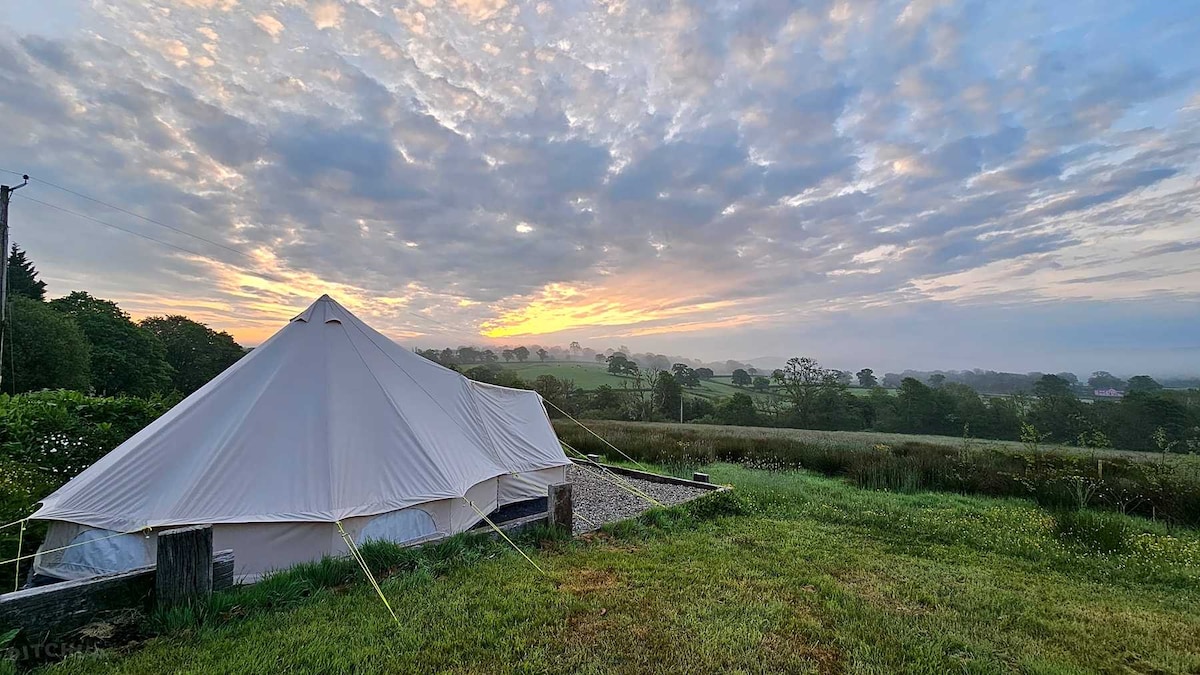 A belle tent is set against a backdrop of rolling greenery, with a cloudy sky tinted by the soft hues of sunrise. The tent is positioned on a grassy area, providing a sense of openness and tranquility.