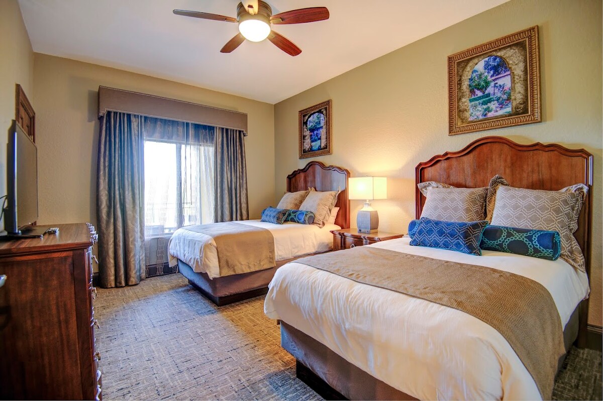 A brightly lit bedroom features two spacious beds, each adorned with coordinating decorative pillows. A wooden dresser and a television are positioned against one wall. Natural light enters through the window dressed with soft curtains, while artwork hangs on the walls, adding character to the space.