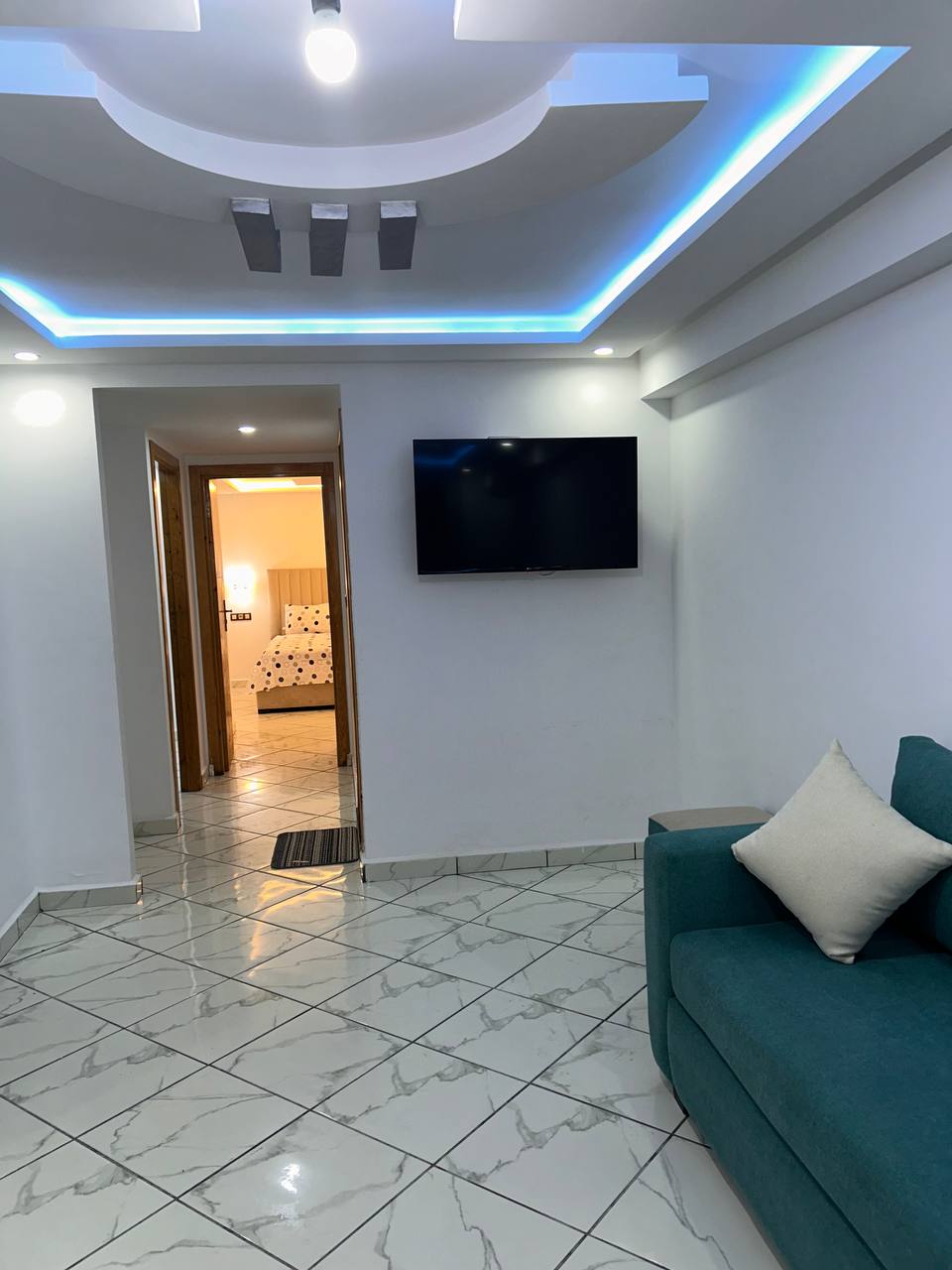 A living area is featured with a turquoise sofa positioned against a white wall. A flat-screen TV is mounted above a small table. The room showcases a modern ceiling with blue LED lights. A doorway leads to a softly lit bedroom visible in the background.