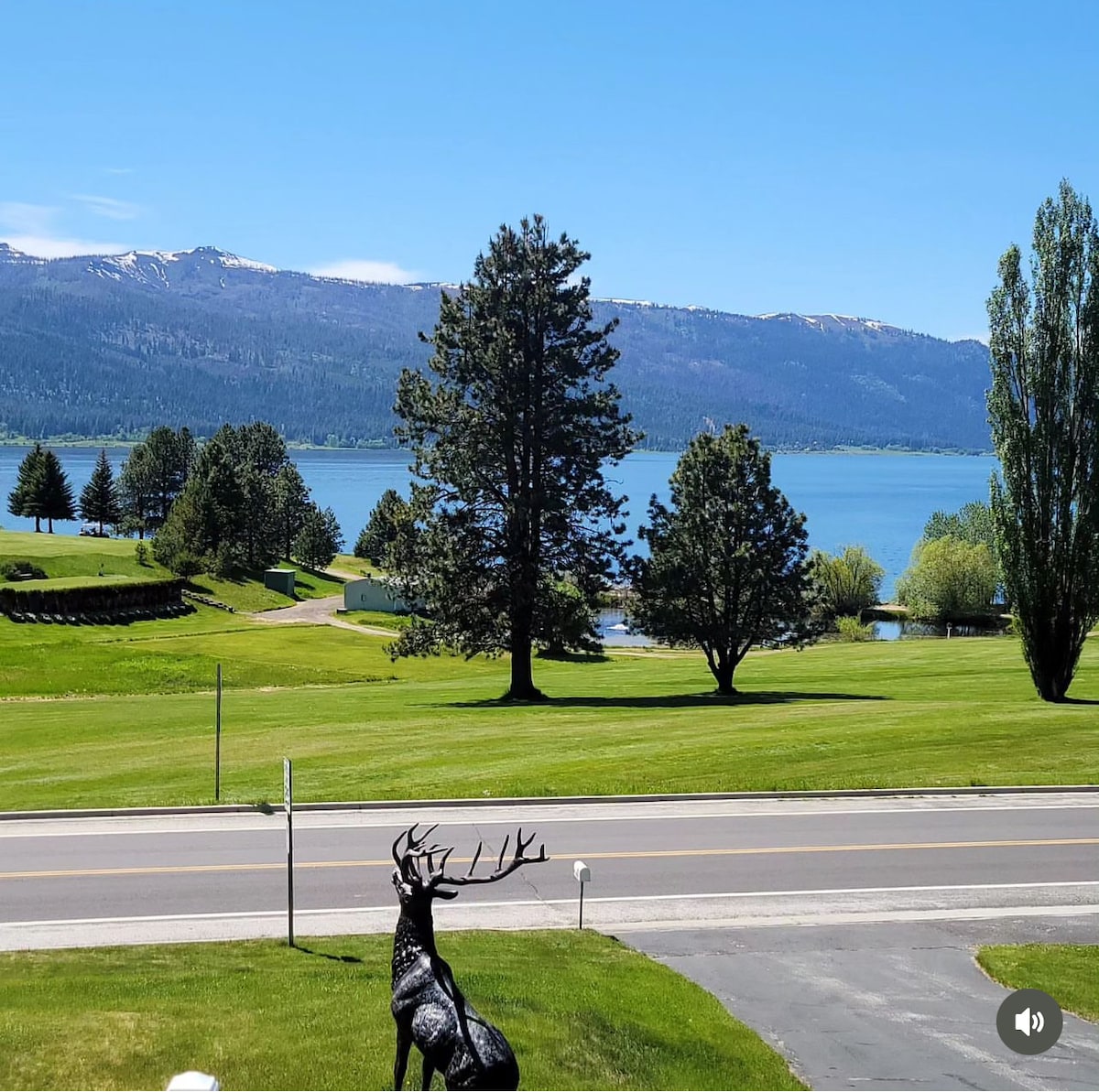 A scenic view of Cascade Lake is presented, framed by lush green lawns and majestic trees. The landscape features distant mountains under a clear blue sky. A black deer sculpture stands in the foreground, adding an artistic touch to the natural surroundings.