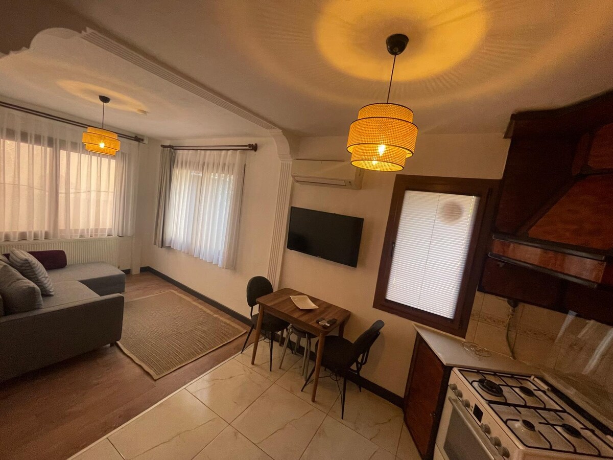 A well-lit living area features large windows allowing natural light to enhance the space. A comfortable gray sofa is positioned alongside a small dining table with two chairs. A mounted television sits on the wall, and stylish pendant lights hang from the ceiling.