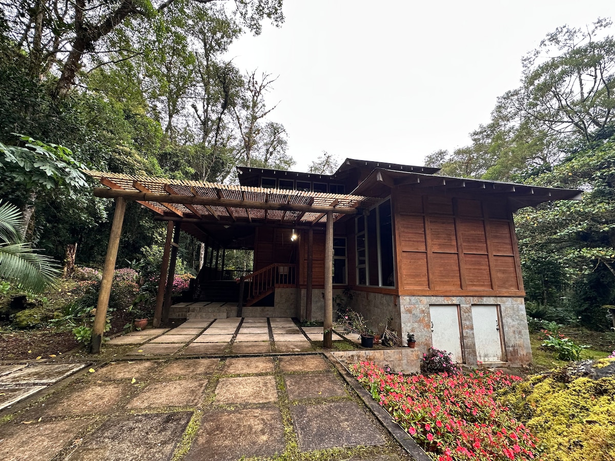 The exterior of the retreat features a wooden structure set against a lush backdrop of trees. A stone pathway leads to the entrance, which is adorned with colorful flowers. A covered porch provides shaded outdoor space, enhancing the connection with nature.