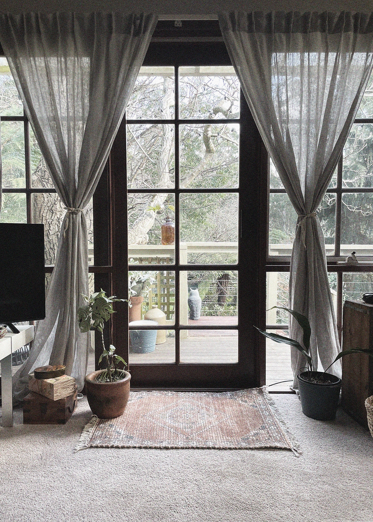 Large windows framed by light grey curtains provide a view of the outdoor deck and surrounding natural greenery. A woven area rug is positioned on the floor, while potted plants add a touch of greenery to the indoor space.