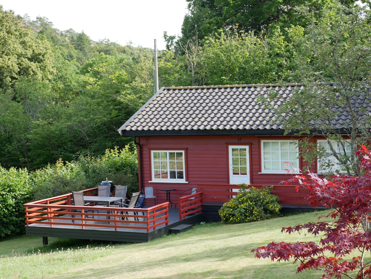 A charming red cabin is set amidst lush greenery, featuring a spacious outdoor deck furnished with a dining table and chairs. A small flowerbed adds a pop of color in front of the cabin, which is complemented by a traditional tiled roof.