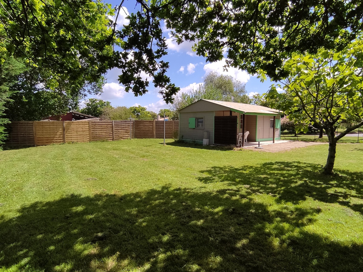 A wooden chalet is situated within a spacious, grassy garden surrounded by trees. The fence provides privacy and the sky is partly cloudy, creating a serene environment. The chalet features windows allowing views of the lush greenery and the natural surroundings.