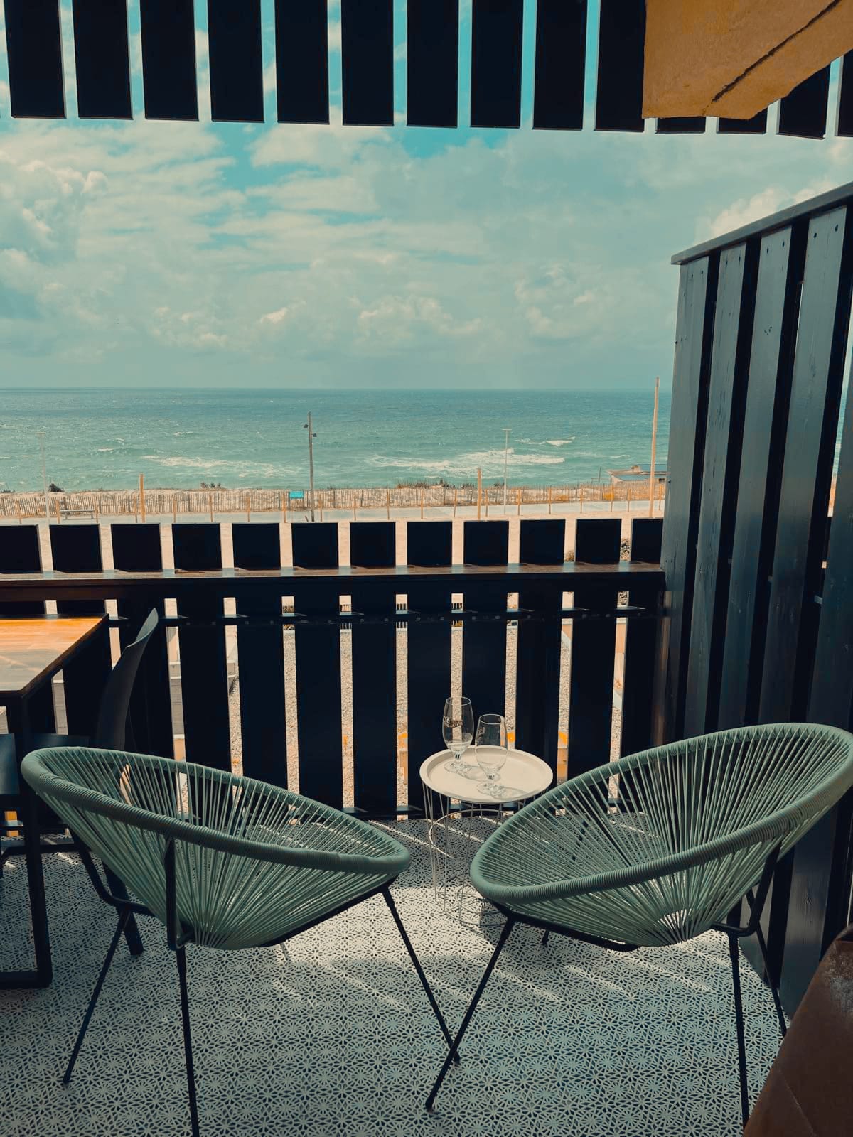 A balcony features two green woven chairs and a small circular table, overlooking the ocean. The serene sea can be seen in the distance, with a partial view of the sky filled with soft clouds. The setting provides a peaceful space for relaxation.