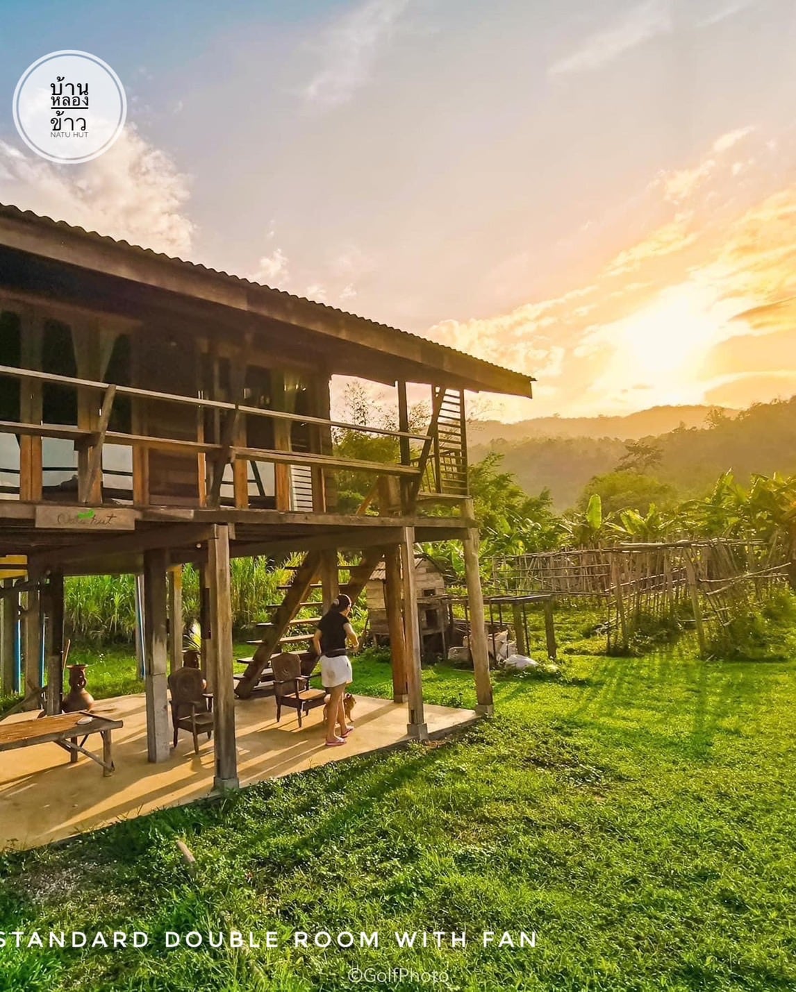 A standard double room constructed with natural materials is set against a backdrop of lush greenery. The warm glow of sunset is visible, and outdoor seating is arranged on the patio. A serene atmosphere is enhanced by the surrounding hills.