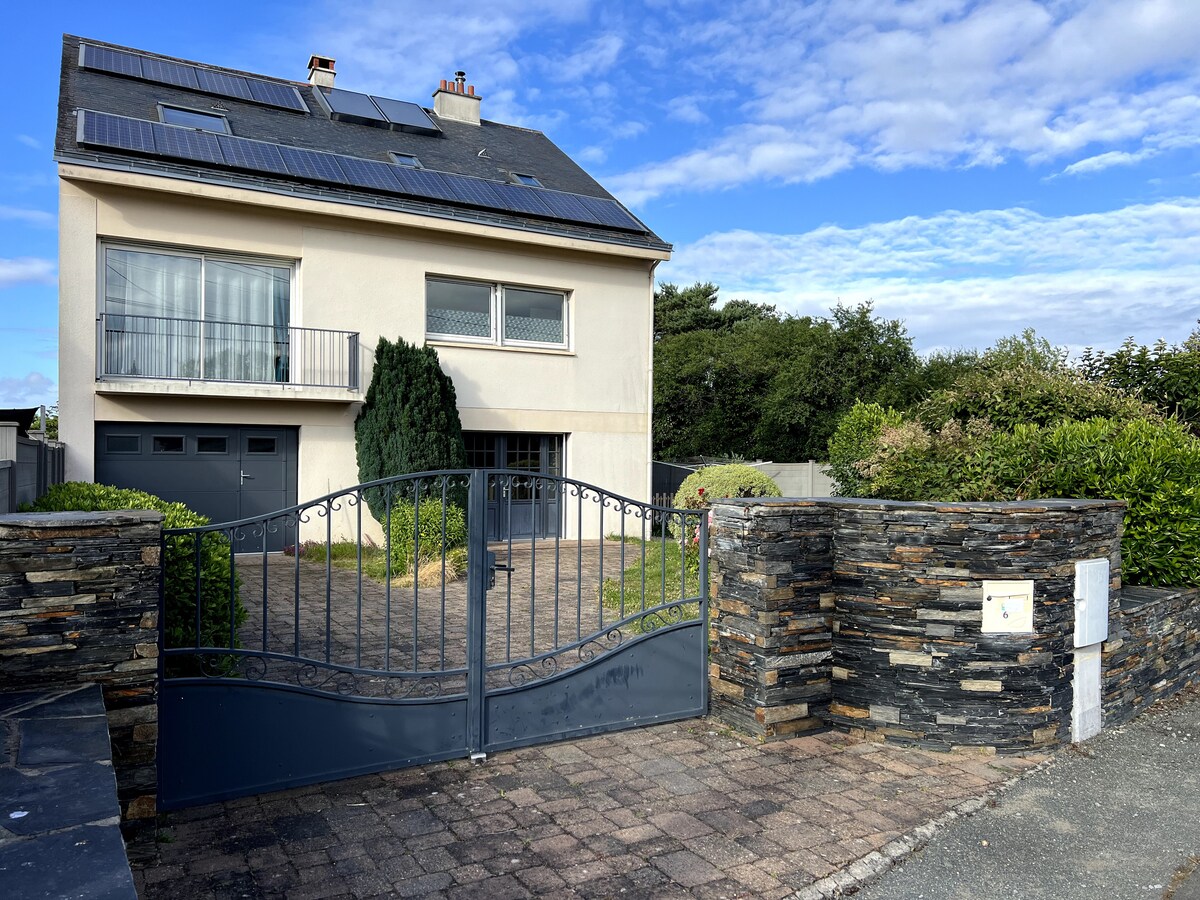 A modern two-story house is set behind a stylish stone wall and gate, with solar panels on the roof. A paved driveway leads to the garage, while well-maintained greenery surrounds the property under a partly cloudy sky.