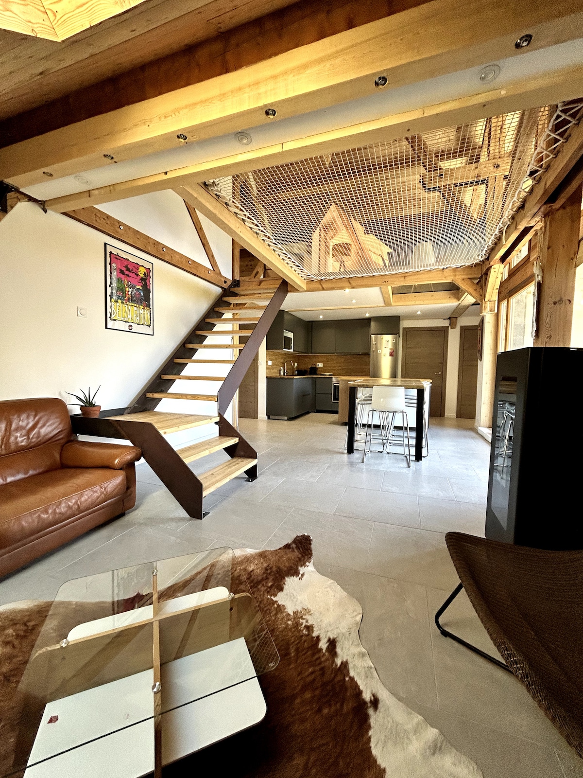 The interior showcases a spacious living area with natural wood beams and a staircase leading to a loft. A brown leather sofa faces a coffee table, while a modern kitchen and dining space are visible in the background. Sleek countertops and ample lighting create a welcoming setting.
