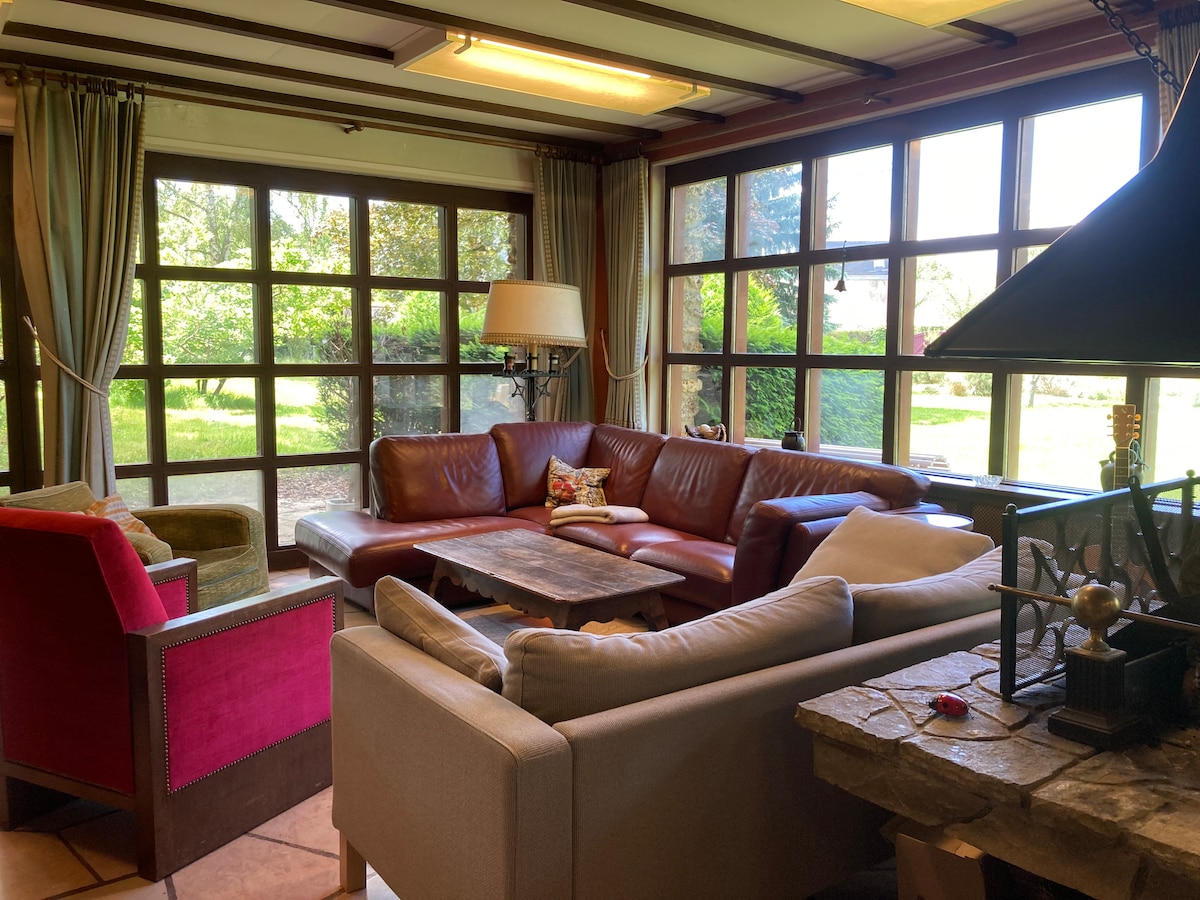 A spacious living area features a large leather sectional sofa and a cozy armchair, accompanied by a decorative table. Natural light floods the room through expansive windows, offering views of the verdant garden outside. A stone fireplace adds a rustic touch to the ambiance.