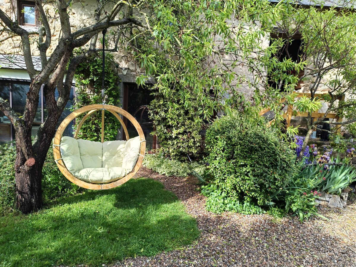 A circular wooden swing with a soft cushion is suspended from a tree branch, surrounded by lush greenery and colorful flowers. The cozy garden area features a well-maintained lawn, inviting relaxation amidst nature.