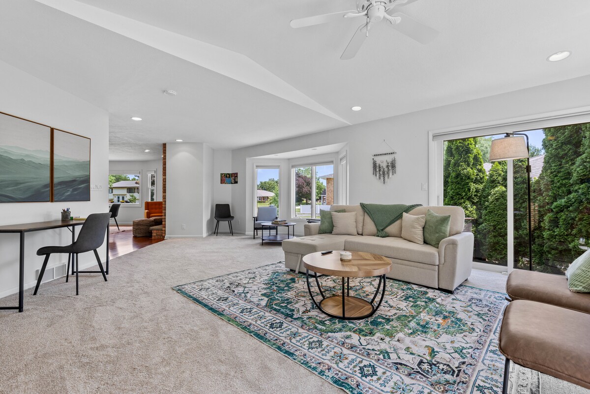 A spacious family room is highlighted by a large sectional sofa and a round coffee table, surrounded by a plush area rug. Natural light enters through glass doors leading to the private deck, while a small dining table is set against one wall.