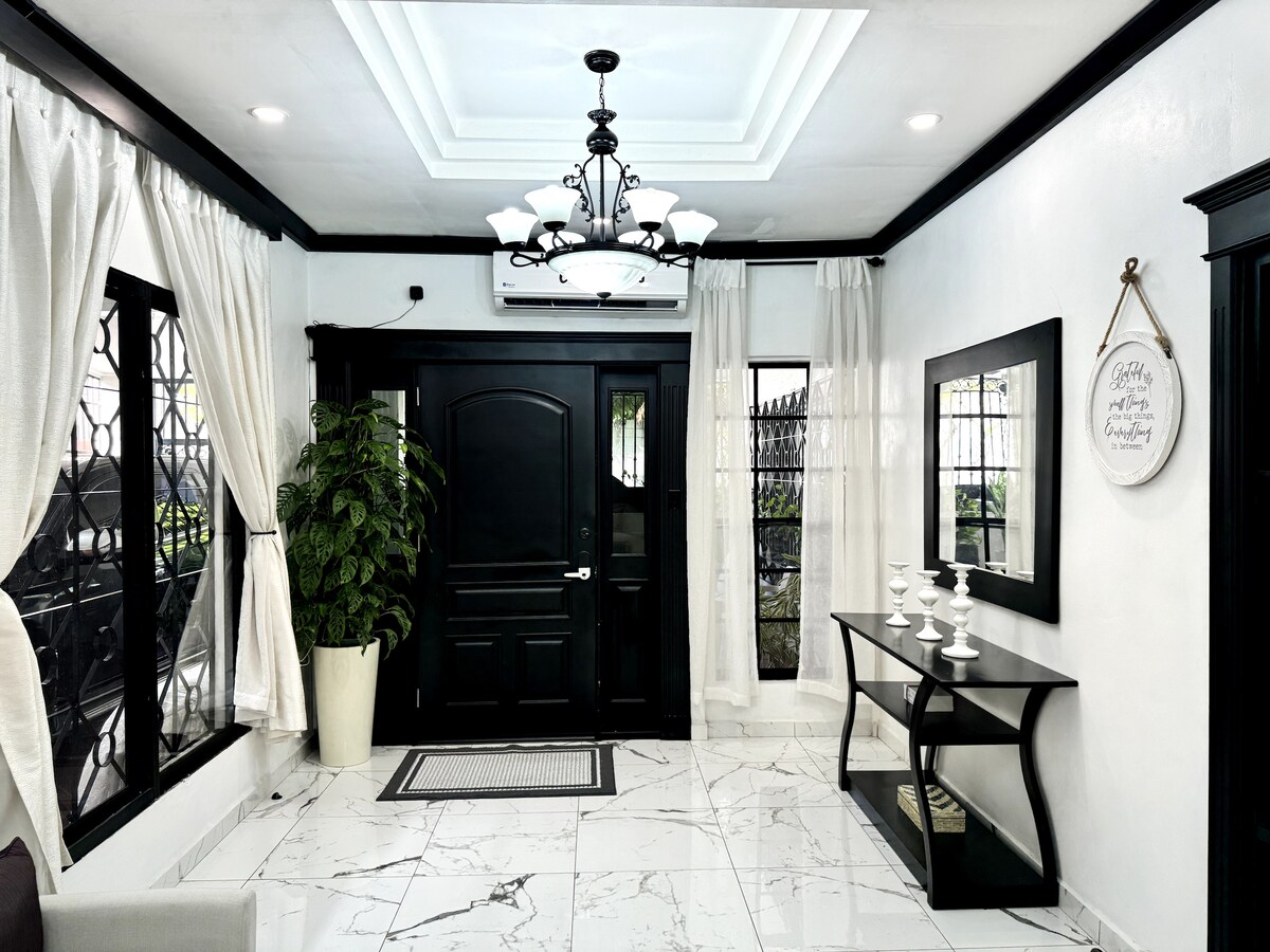 The entryway features a black door framed by large windows covered with sheer curtains, allowing soft light to filter through. A stylish chandelier hangs above a marble floor. A sleek table displays decorative items, while a large potted plant adds a touch of greenery.