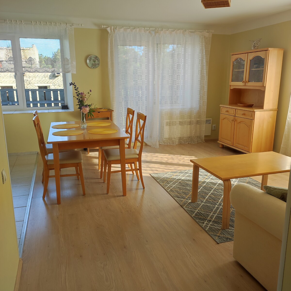 A bright living and dining area is featured, with a wooden table set for four and matching chairs. Natural light filters through sheer curtains covering the windows. A light-colored sofa and a wooden cabinet add warmth, complemented by a patterned area rug.
